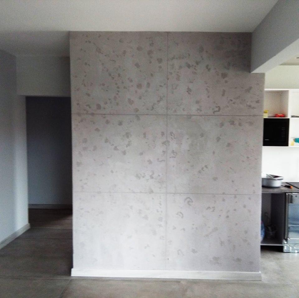 Ściana wykończona betonem architektonicznym w jasnoszarym odcieniu, podzielona na sześć kwadratowych płyt z widocznymi fugami, w tle fragment kuchni i korytarza.