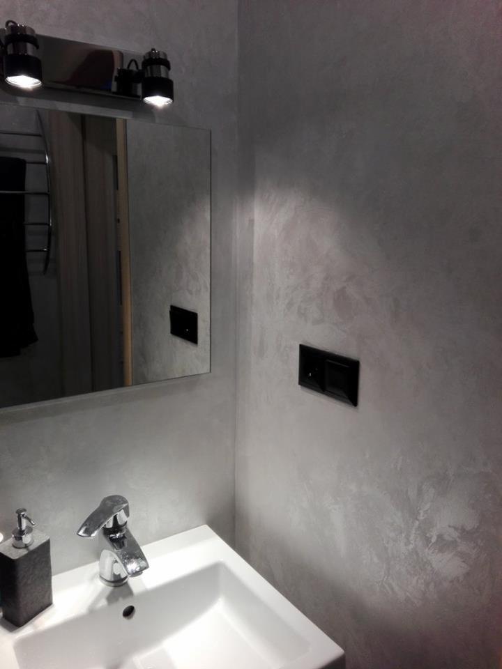 Ściana w łazience z dekoracyjną, szarą fakturą, umywalka z baterią i dozownikiem mydła w kolorze grafitowym, czarny włącznik światła na ścianie, lustro z oświetleniem punktowym.