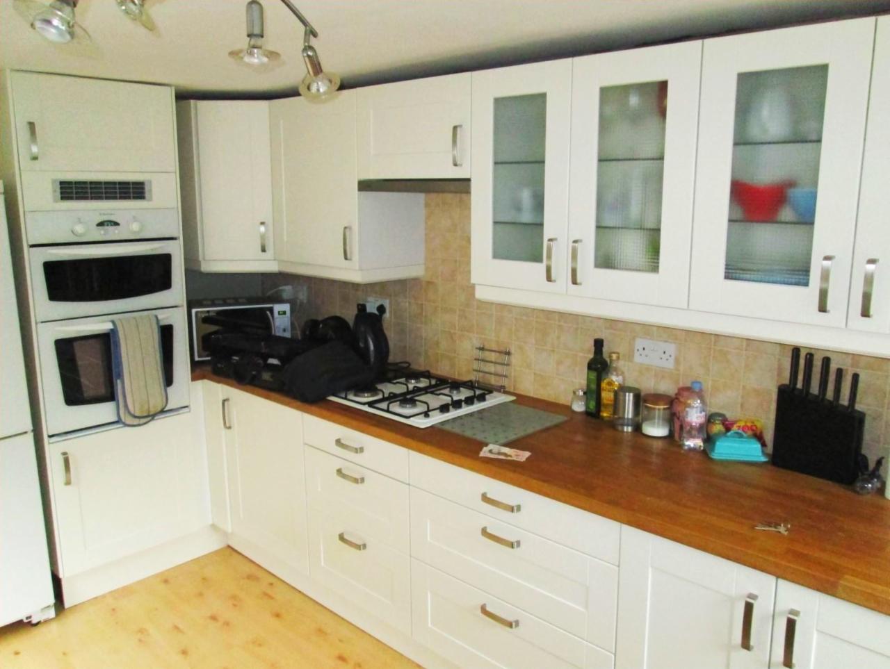 Jasna kuchnia z drewnianym blatem, białymi szafkami z przeszklonymi frontami i sprzętem AGD; widoczny piec, płyta gazowa i drobne akcesoria kuchenne.