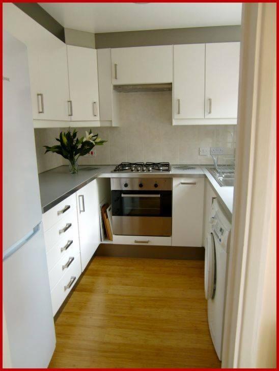 Jasna kuchnia w bloku z białymi szafkami, drewnianą podłogą i bukietem białych kwiatów na blacie, widoczna pralka i piekarnik.