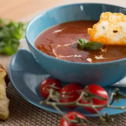 Aromatyczna zupa pomidorowa w turkusowej misce, udekorowana kleksem śmietany, listkiem bazylii i chrupiącym chips z sera, podana z ziołowym pieczywem i gałązką pomidorków cherry.