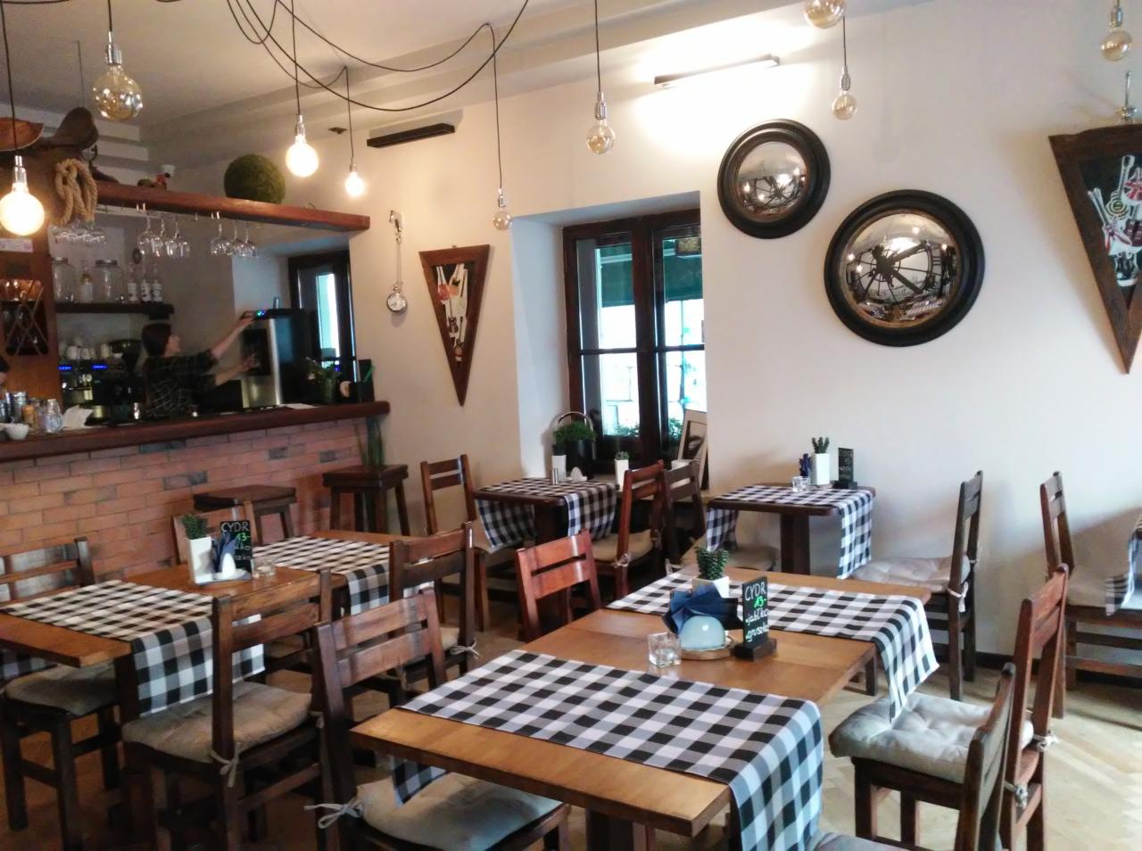 Wnętrze kawiarni lub restauracji z drewnianymi stołami nakrytymi obrusami w czarno-białą kratę, ceglanym barem, dekoracyjnymi lustrami i żarówkami Edison