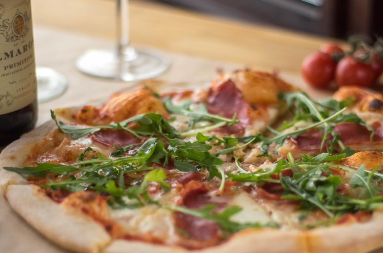 Pizza z rukolą i salami na drewnianym stole, obok butelka wina i pomidory koktajlowe.