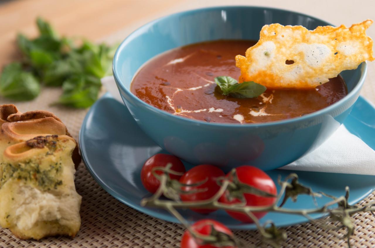 Aromatyczna zupa pomidorowa w turkusowej misce, udekorowana kleksem śmietany, listkiem bazylii i chrupiącym chips z sera, podana z ziołowym pieczywem i gałązką pomidorków cherry.