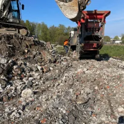 Ciężki sprzęt budowlany, w tym koparka i kruszarka, pracują na placu budowy, gdzie gruz i resztki cegieł tworzą stosy; pracownik w pomarańczowej kamizelce nadzoruje proces.