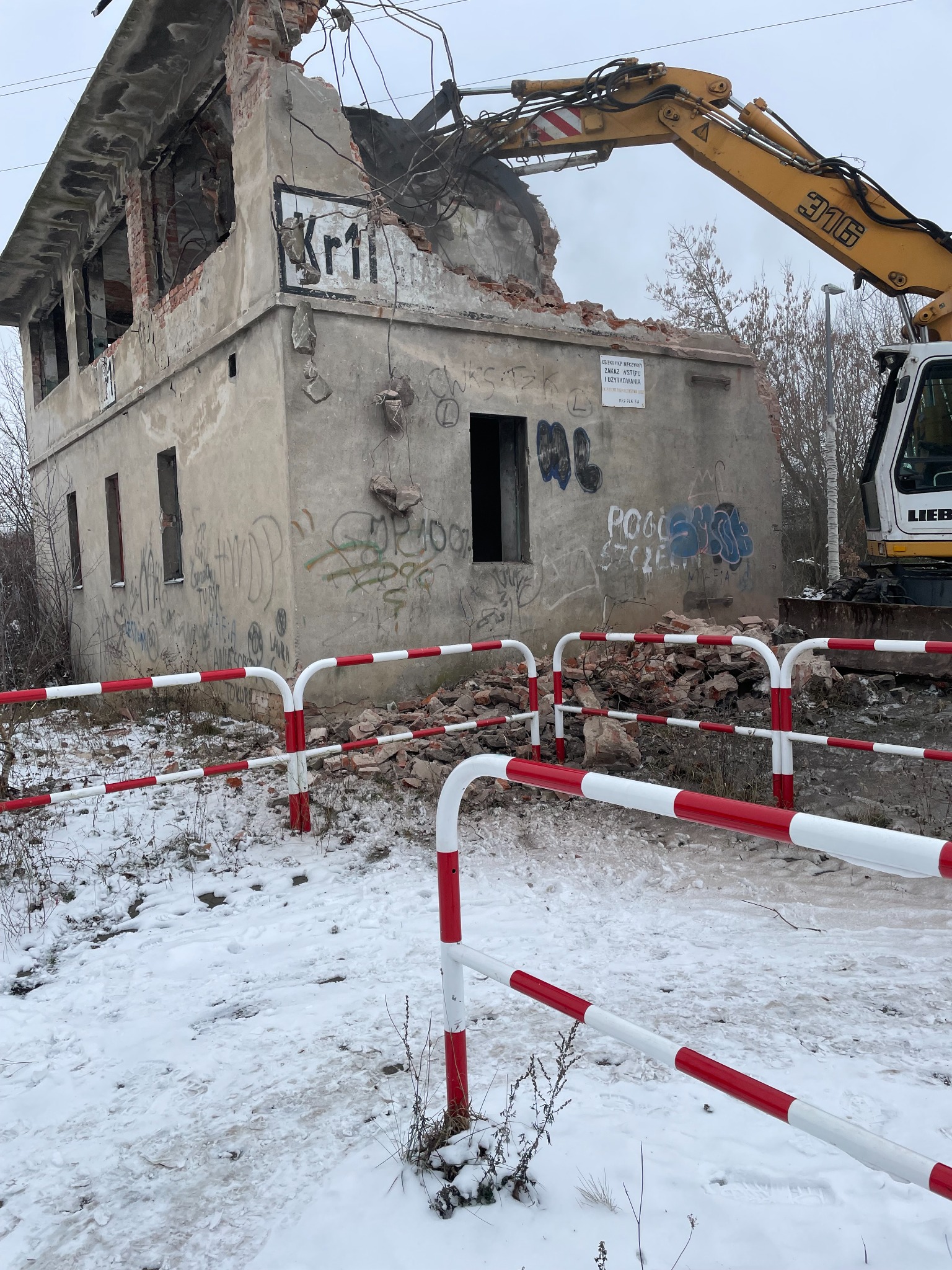Żółta koparka Liebherr 316 w trakcie demontażu dwukondygnacyjnego, betonowego budynku pokrytego graffiti, zabezpieczonego barierami ochronnymi w biało-czerwone pasy na ośnieżonym terenie.