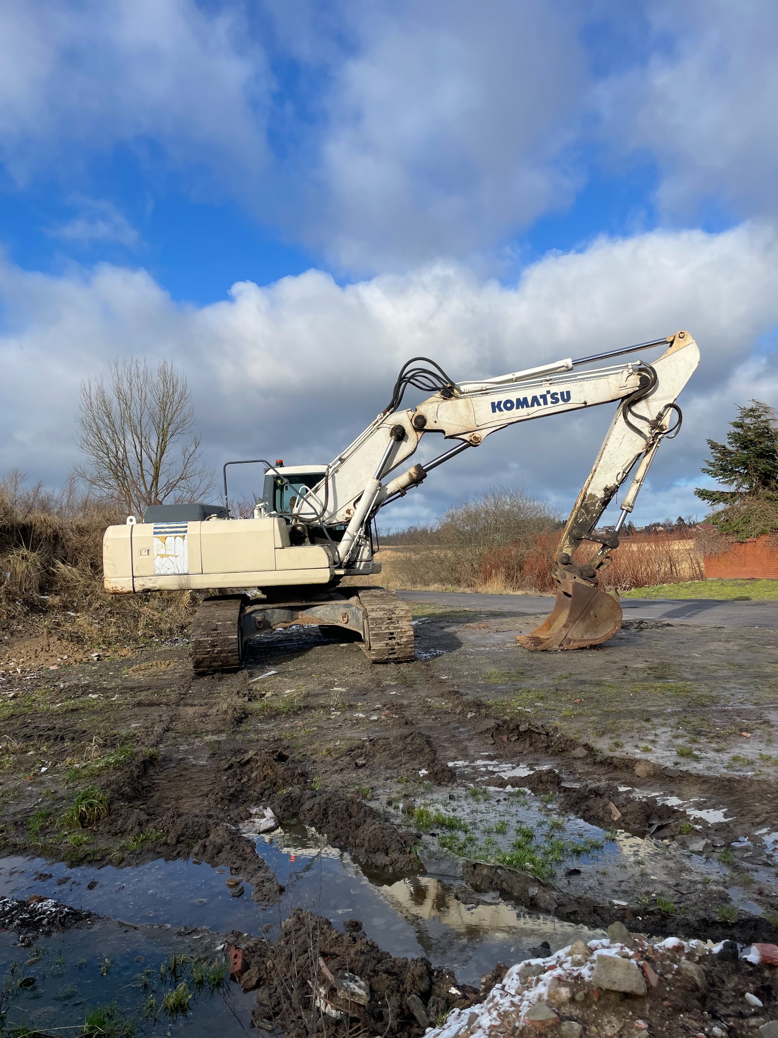 Biała koparka Komatsu stoi na błotnistym terenie z kałużami, w tle błękitne niebo z chmurami.