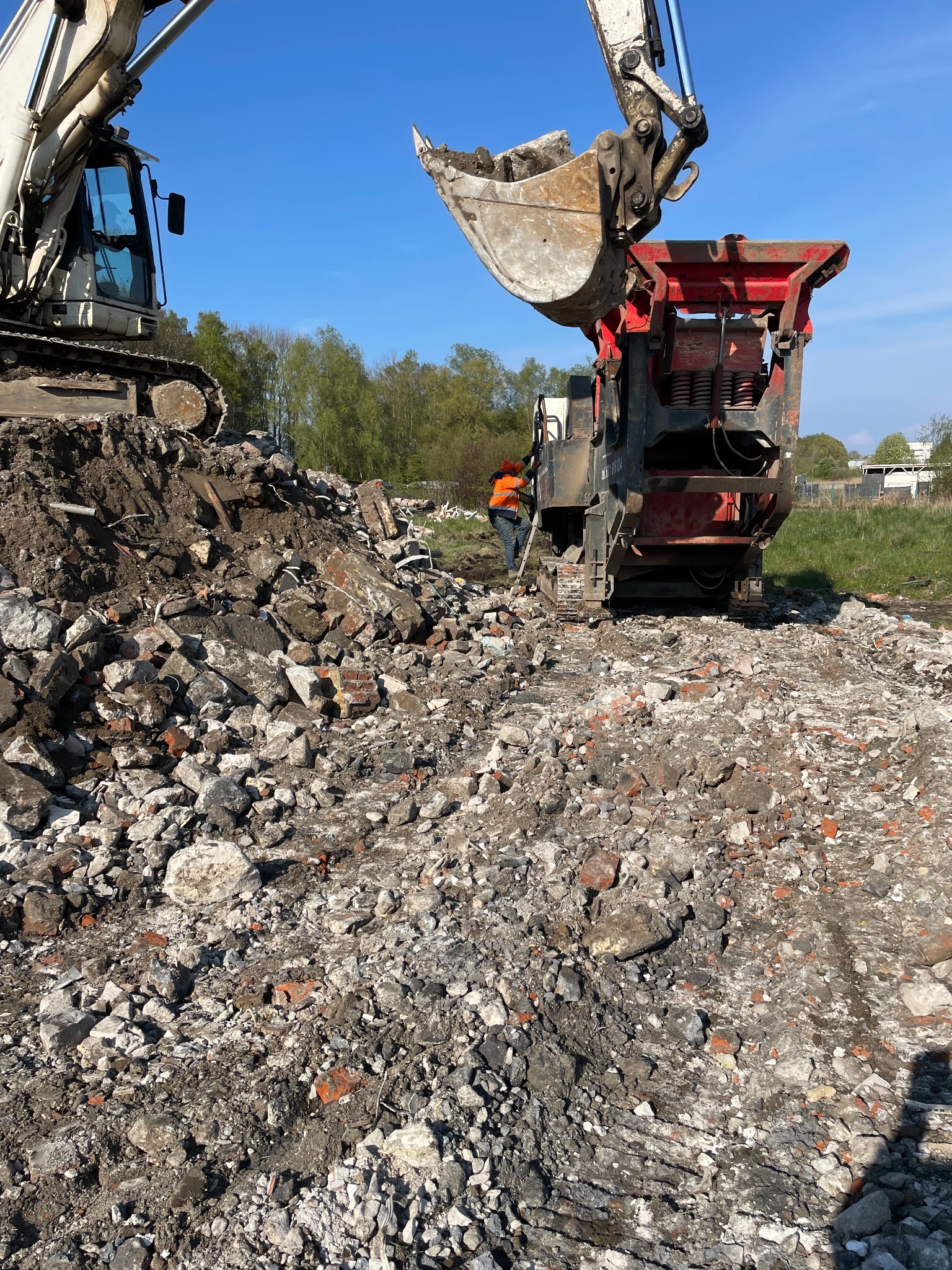 Ciężki sprzęt budowlany, w tym koparka i kruszarka, pracują na placu budowy, gdzie gruz i resztki cegieł tworzą stosy; pracownik w pomarańczowej kamizelce nadzoruje proces.