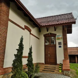 Budynek z białymi ścianami i ceglanymi detalami, brązowe drzwi wejściowe z tabliczką 'Bajka', schody z szarej kostki, małe drzewka iglaste przy ścianie, pochmurne niebo.