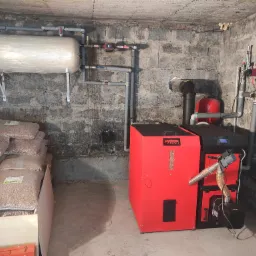 Czerwony piec na pellet z podajnikiem, obok zapas pelletu w workach, instalacja hydrauliczna z zbiornikiem wyrównawczym w piwnicy o ścianach z szarego betonu.