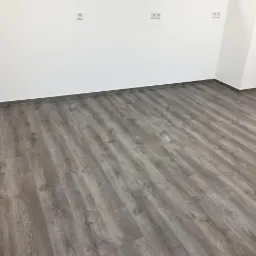 Szare panele podłogowe imitujące drewno ułożone w pustym, jasnym pomieszczeniu z białymi ścianami i kilkoma gniazdkami elektrycznymi.
