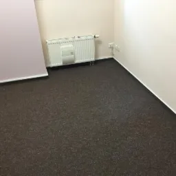 Ciemnobrązowa, teksturowana podłoga w pokoju z grzejnikiem i gniazdkami elektrycznymi przy ścianie.