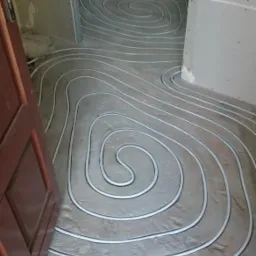 Wnętrze w trakcie montażu ogrzewania podłogowego z widocznymi pętlami rur ułożonymi na szarej wylewce, częściowo zakryte folią ochronną w tle.