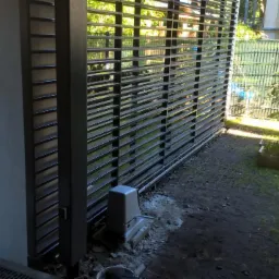 Nowoczesna brama przesuwna z automatyką w trakcie montażu, widoczny siłownik, betonowy słupek i fragment trawnika z skrzynką na rośliny.