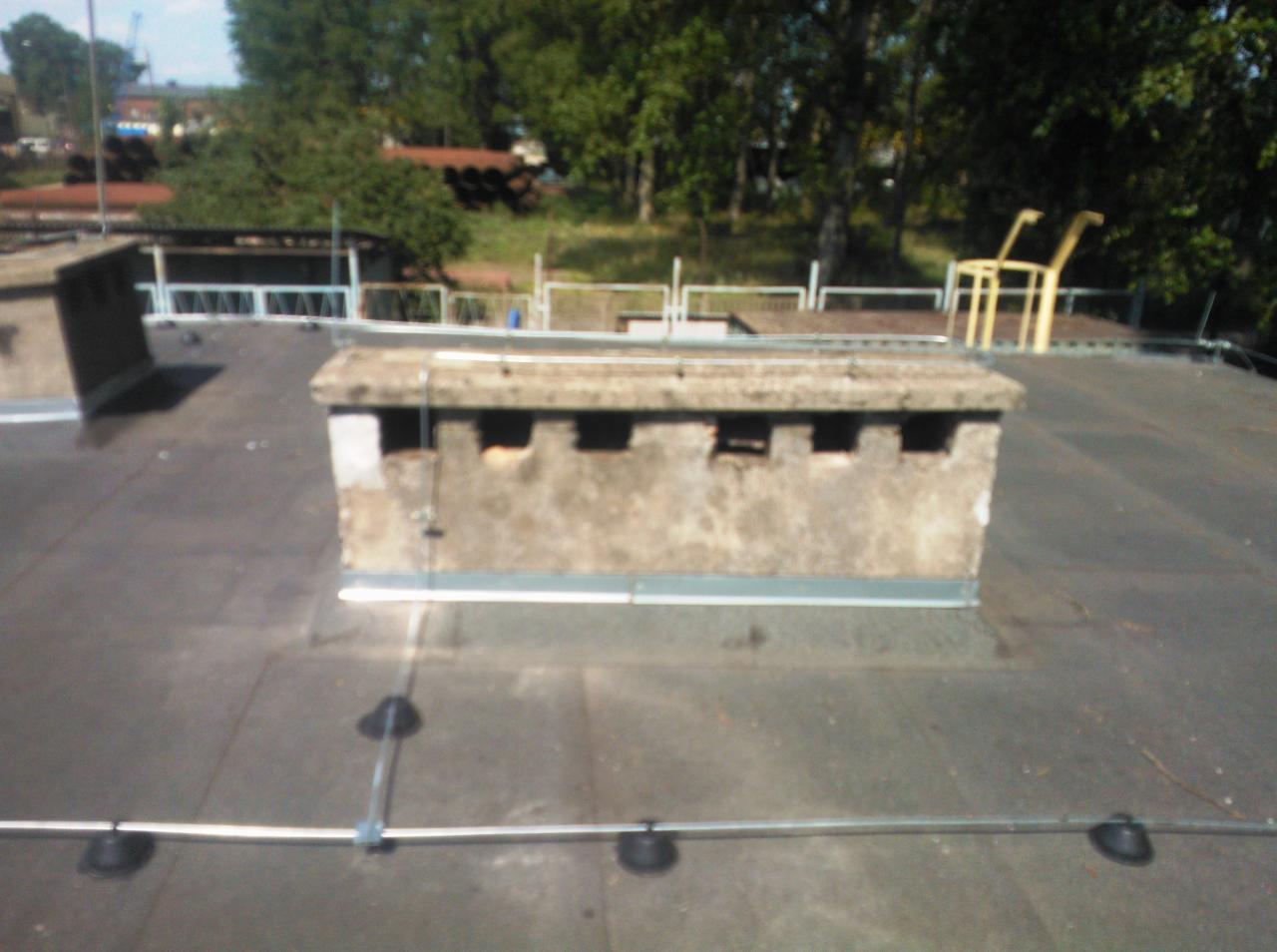 Betonowy komin z otworami na dachu pokrytym papą, zabezpieczony metalowym uziomem.