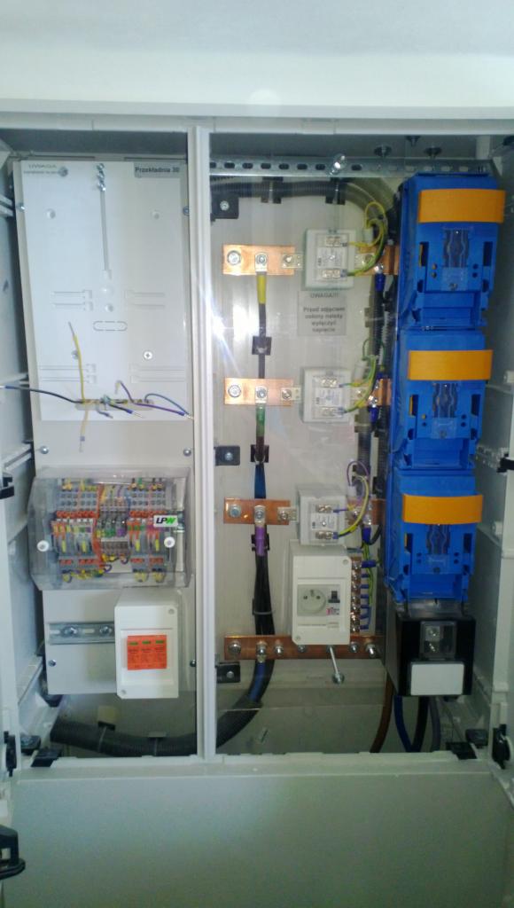 Otwarta skrzynka rozdzielcza z widocznymi przewodami, listwami zaciskowymi, bezpiecznikami i innymi elementami instalacji elektrycznej, wewnątrz pomieszczenia.