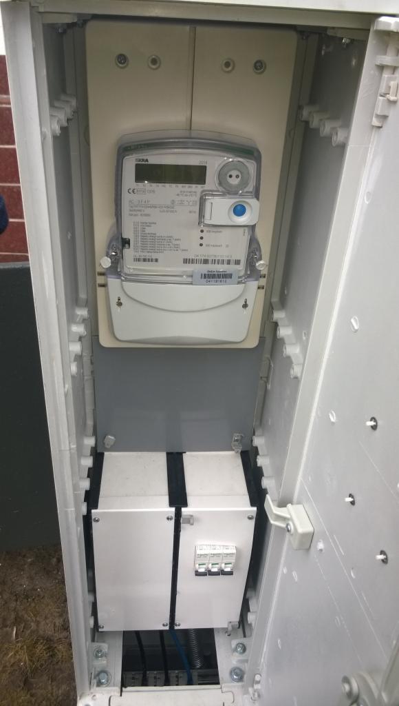 Otwarta skrzynka z licznikiem energii elektrycznej i zabezpieczeniami, widoczne przewody.