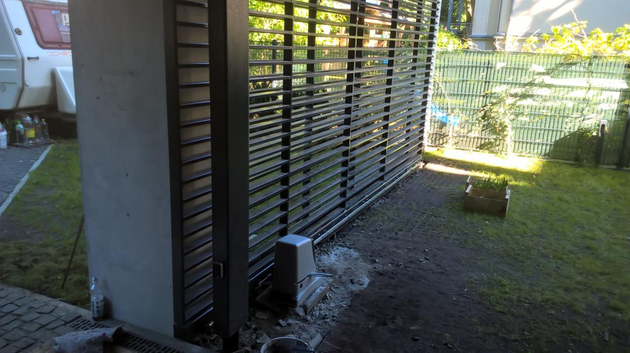 Nowoczesna brama przesuwna z automatyką w trakcie montażu, widoczny siłownik, betonowy słupek i fragment trawnika z skrzynką na rośliny.