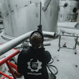 Pracownik w bluzie z logo Weldgam spawa metalową konstrukcję w hali. Widoczne iskry i elementy konstrukcji.