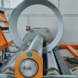 Proces gięcia metalu na walcarce z widocznym elementem w kształcie okręgu. Maszyna w kolorach szarym i pomarańczowym, z naklejką 'Kontrola Jakości'.