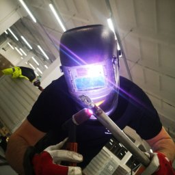 WELD-GAM Szymon Gamalczyk - Spawanie rury metodą TIG w hali. Widoczny spawacz w masce ochronnej, trzymający uchwyt spawalniczy. Intensywne światło łuku spawalniczego. Ujęcie z dołu.