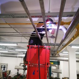 WELD-GAM Szymon Gamalczyk - Spawanie rur w Bożej Woli z podnośnika. Spawacz w kombinezonie i masce spawalniczej łączy metalowe elementy instalacji pod sufitem w hali przemysłowej.