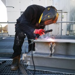 WELD-GAM Szymon Gamalczyk - Spawacz w masce spawalniczej łączy metalowe elementy konstrukcji na zewnątrz budynku. Widoczne iskry i spawarka. Na podłodze leżą elektrody spawalnicze.