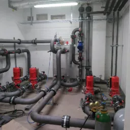 Instalacja przemysłowa z rurami, czerwonymi pompami i zieloną butlą z gazem w pomieszczeniu technicznym.