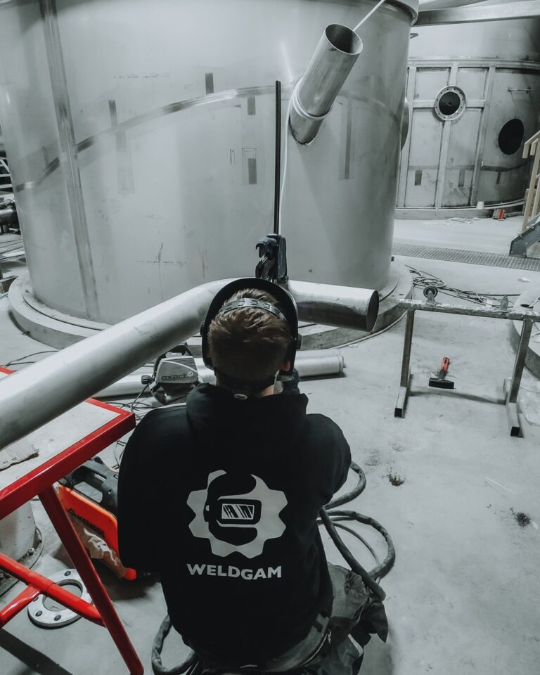 Pracownik w bluzie z logo Weldgam spawa metalową konstrukcję w hali. Widoczne iskry i elementy konstrukcji.