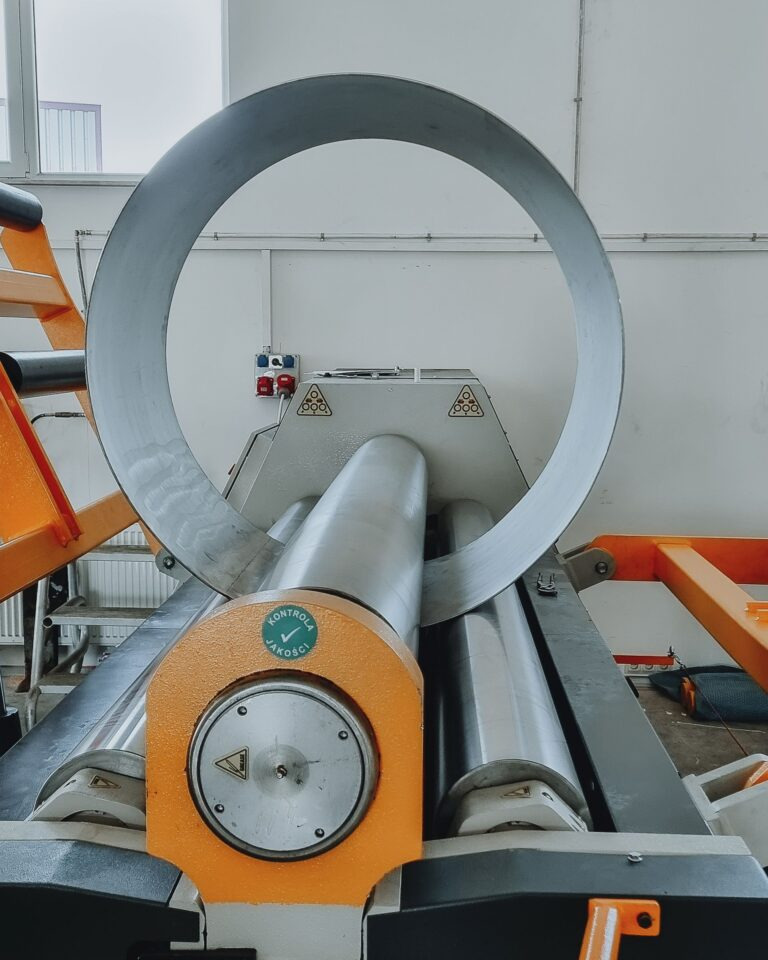 Proces gięcia metalu na walcarce z widocznym elementem w kształcie okręgu. Maszyna w kolorach szarym i pomarańczowym, z naklejką 'Kontrola Jakości'.