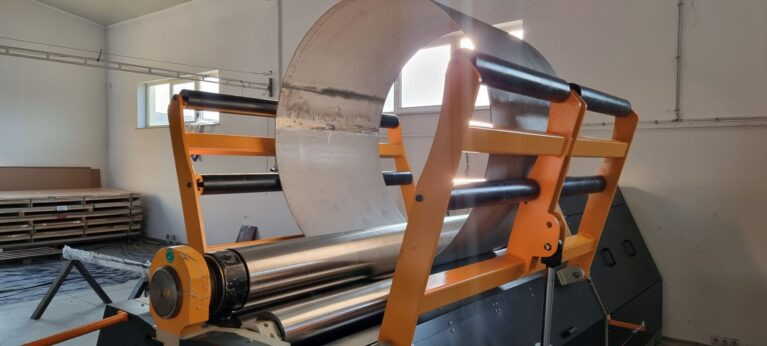 Proces formowania blachy na walcarce z pomarańczową ramą w hali produkcyjnej. Widoczne rolki i częściowo uformowany element. Jasne oświetlenie.