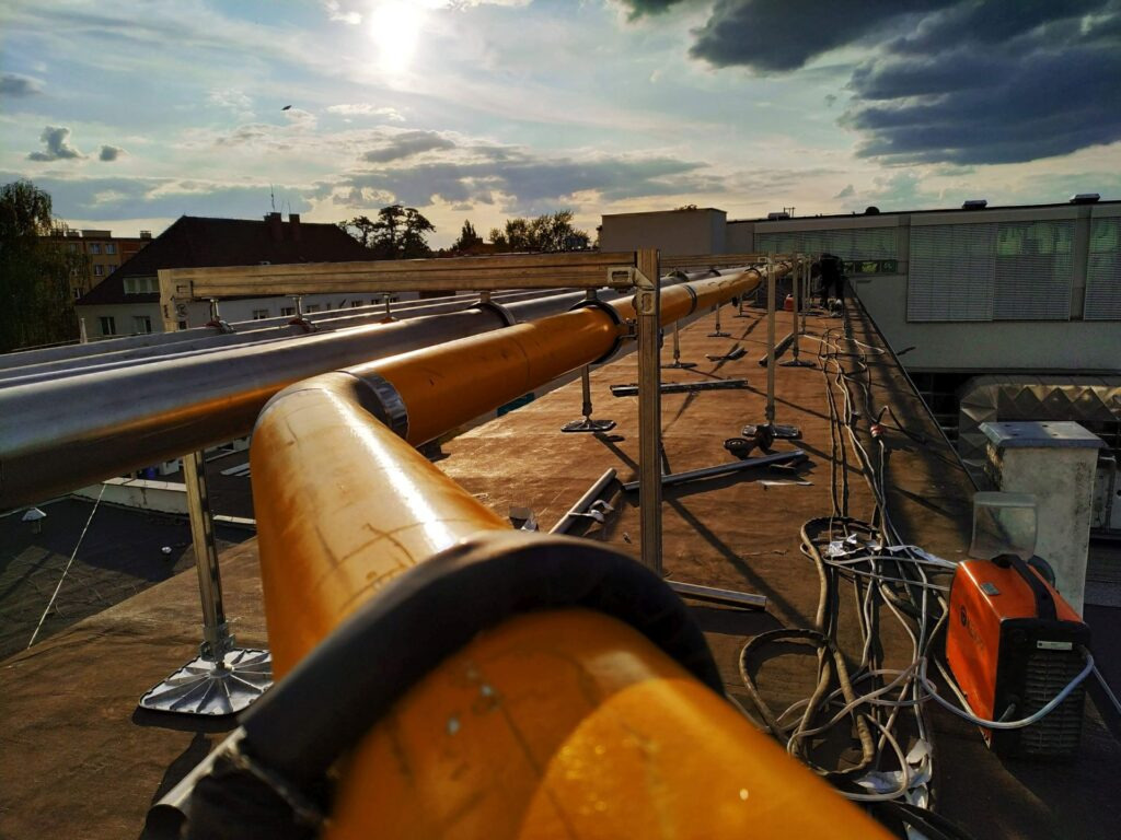 Instalacja przemysłowa z rur na dachu budynku, widoczny sprzęt spawalniczy i pracownik w oddali. Rury w kolorze srebrnym i pomarańczowym, mocowane na metalowych podporach.
