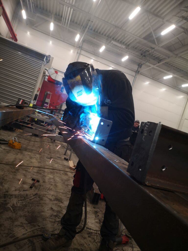 Pracownik w masce spawalniczej łączy metalowe elementy konstrukcji w hali. Widoczne iskry i intensywne światło łuku elektrycznego. W tle brama i częściowo widoczny czerwony samochód.