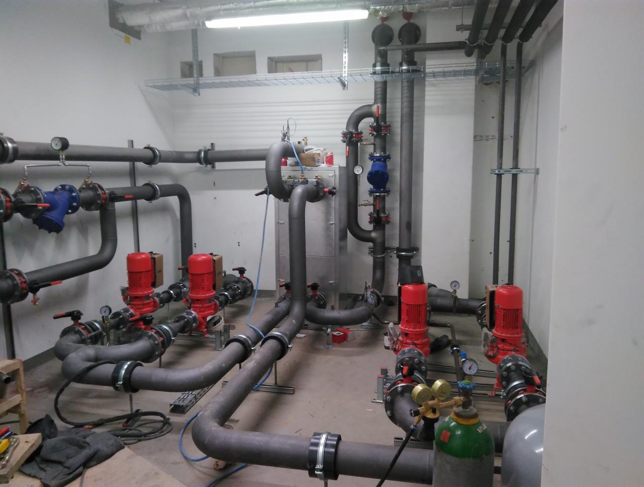 Instalacja przemysłowa z rurami, czerwonymi pompami i zieloną butlą z gazem w pomieszczeniu technicznym.