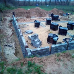 Budowa fundamentów z bloczków betonowych, zbrojenie pionowe wystające z muru, teren przygotowany pod wylewkę, w tle drzewa i hałdy ziemi.