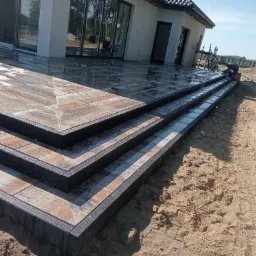 Wykończony taras z płytkami imitującymi drewno, z widocznymi schodami i obrzeżami, z pracującym wykonawcą i zielonym wózkiem widłowym w tle.