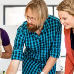 Trzy osoby pochylone nad dokumentem podczas spotkania, uśmiechnięte, w tle biała ściana i okno.