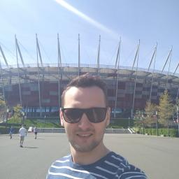 EL-BOL, Usługi Elektryczne, Smart Home - Mężczyzna w okularach na tle Stadionu Narodowego w słoneczny dzień. W tle spacerujący ludzie. Ujęcie z dołu, podkreślające architekturę stadionu.