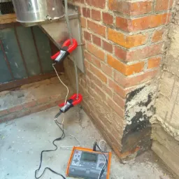 Pomiar rezystancji uziemienia za pomocą miernika cęgowego i urządzenia pomiarowego przy ścianie z czerwonej cegły z widocznym kominem.