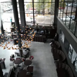 Wysoki hol restauracji z widokiem z góry na stoliki, krzesła i bar, oświetlony dekoracyjnym żyrandolem w kształcie kuli z licznymi punktami świetlnymi, duże okna z widokiem na ulicę.