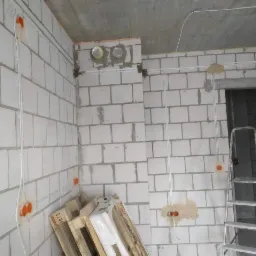 Surowy stan wnętrza z widocznymi przewodami elektrycznymi prowadzonymi po ścianach z betonu komórkowego, puszki instalacyjne w kolorze pomarańczowym, drabina oparta o ścianę, palety na podłodze.