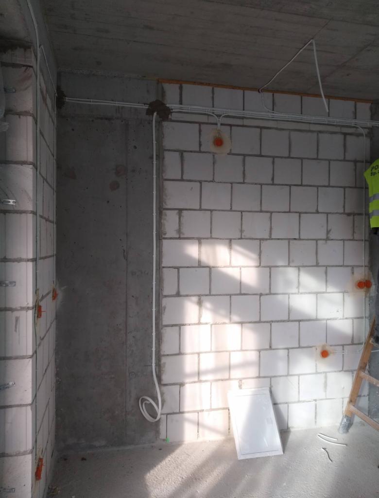 Surowy stan wnętrza w trakcie prac: białe kable poprowadzone po ścianach z bloczków, widoczne puszki instalacyjne, betonowy sufit, fragment drabiny i osoba w odblaskowej kamizelce w tle.