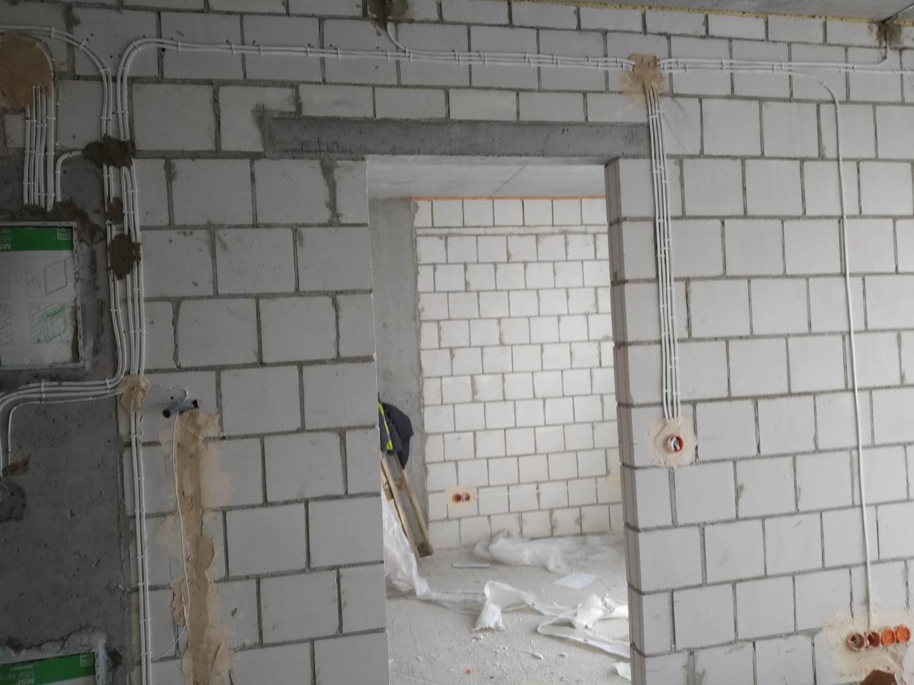 Surowe ściany z betonu komórkowego w trakcie rozprowadzania przewodów elektrycznych; widoczne puszki instalacyjne i kable prowadzone natynkowo, zabezpieczone uchwytami; w tle widoczne kolejne...