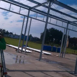 Człowiek w zielonej bluzie mierzy kąty za pomocą niwelatora na tle konstrukcji stalowej hali w budowie, widoczne niebo i teren zielony.