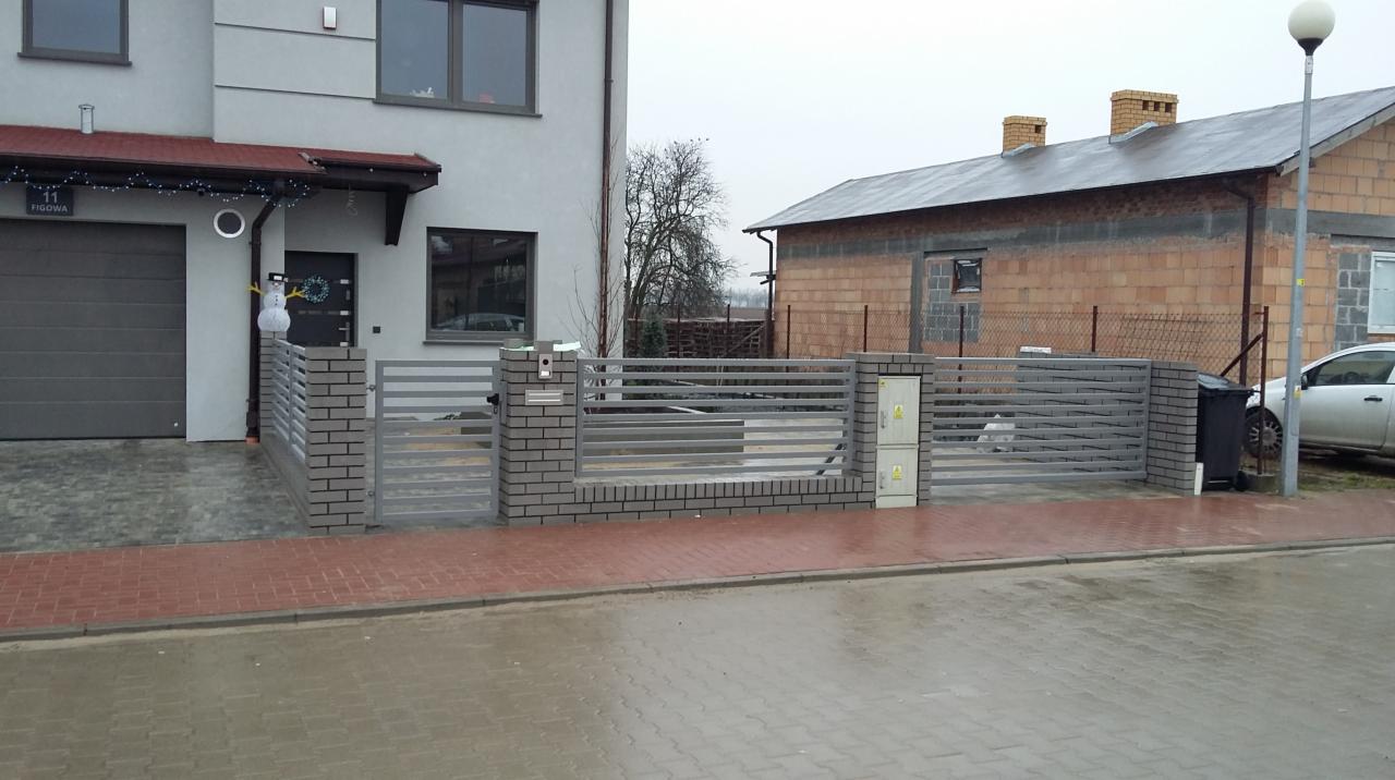 Nowoczesne ogrodzenie posesji z szarych, poziomych przęseł zamontowanych między słupkami z szarej cegły klinkierowej, z widoczną skrzynką elektryczną na jednym ze słupków, na tle domu z szarą...
