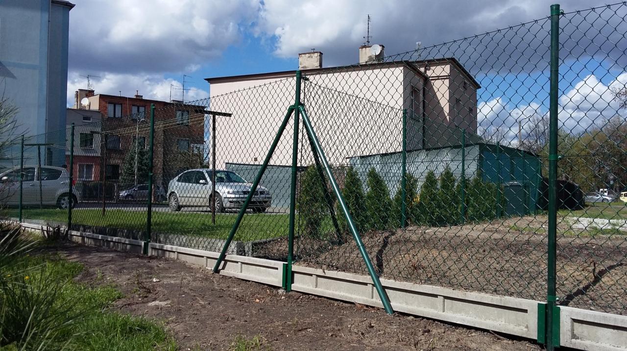 Nowo postawione ogrodzenie z siatki z zielonymi słupkami na betonowej podmurówce na tle budynków mieszkalnych i samochodów.