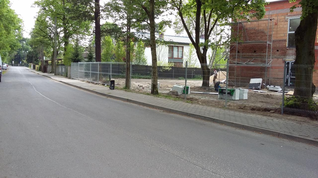 Widok z ulicy na montaż ogrodzenia z siatki metalowej na posesji z budynkiem w trakcie budowy, widoczne materiały budowlane i pracownik w oddali.