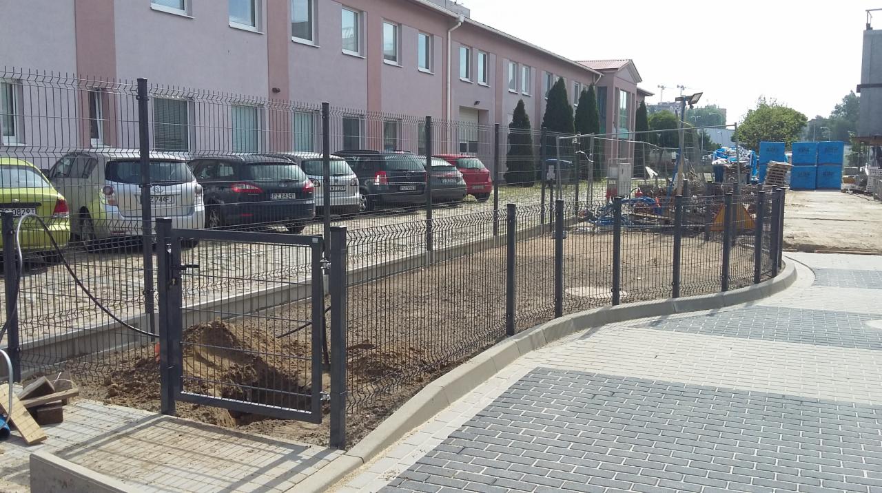 Montaż grafitowego ogrodzenia panelowego z furtką na terenie przemysłowym, widoczne zaparkowane samochody oraz materiały budowlane.