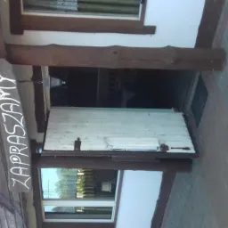 Wejście do lokalu z drewnianym szyldem 'Zapraszamy' nad drzwiami, widok z zewnątrz.