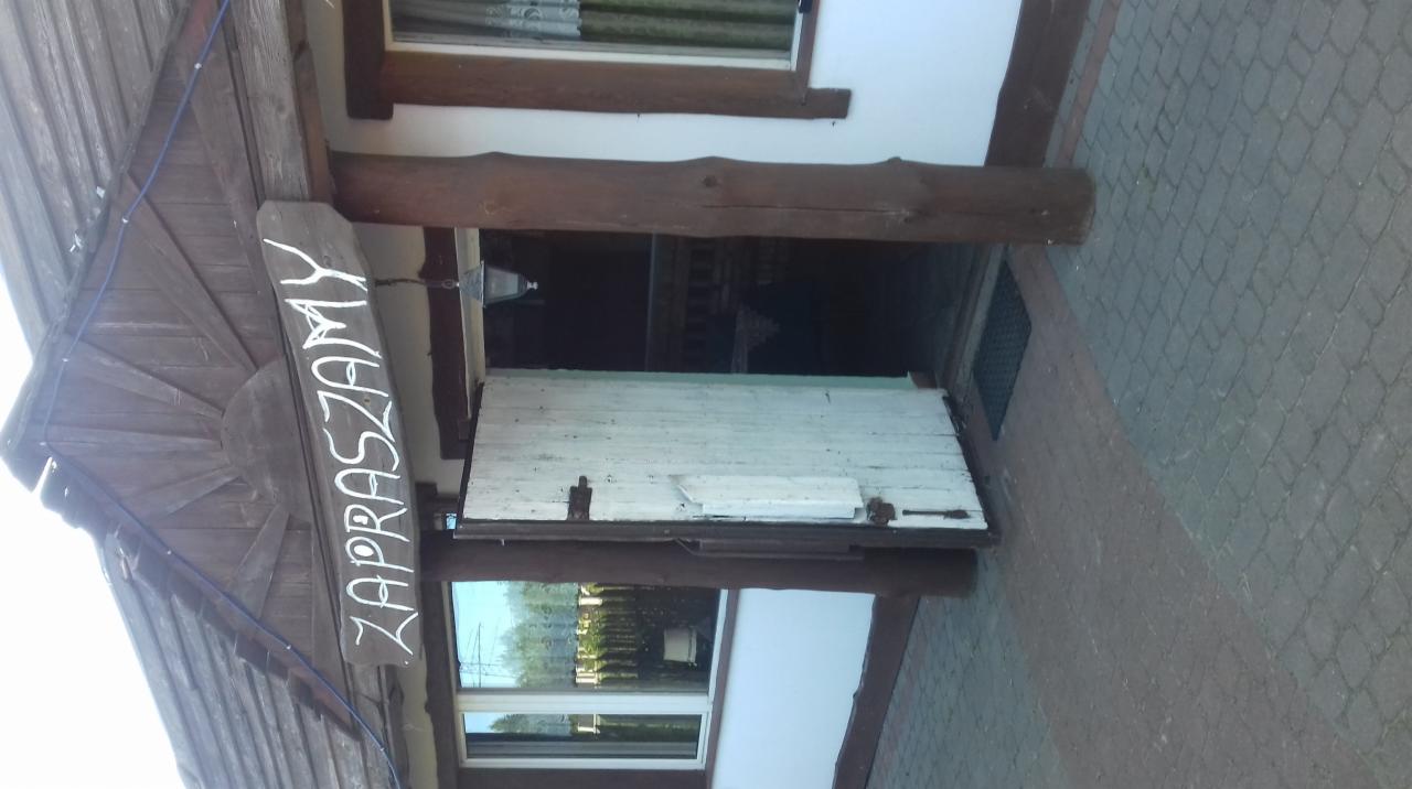 Wejście do lokalu z drewnianym szyldem 'Zapraszamy' nad drzwiami, widok z zewnątrz.